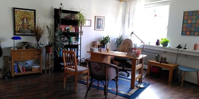 Praxisraum mit Schreibtisch, einem Stuhl und einem Rattansessel, Bücherregale, Bilder ander Wand.